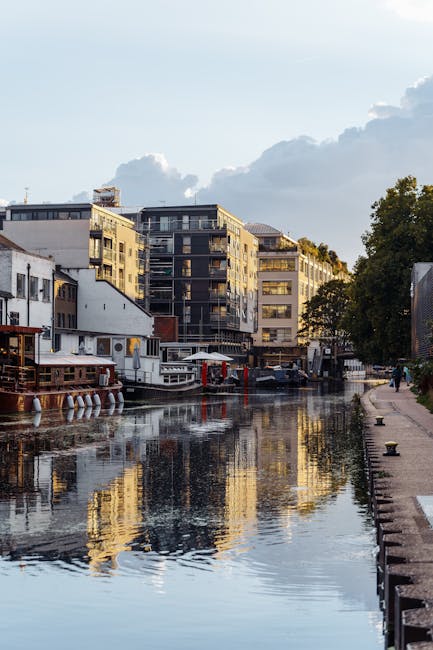 A panoramic view of a canal lined with modern multi-storey residential buildings and boats docked along the water's edge. The buildings feature large windows and balconies, with some balconies having glass railings. On the left side, there are structures with white facades and wooden elements, possibly houseboats or floating restaurants, with outdoor seating areas and awnings visible. The canal's calm water reflects the buildings, boats, and partly cloudy sky above, creating mirrored images. On the right side, a stone-paved walkway runs parallel to the canal, with a few trees providing greenery. In the foreground, a section of the pavement includes a bollard and a mooring post. The scene is illuminated by natural daylight, suggesting a clear, mild day. This setting exemplifies urban house removals and furniture transport processes at a residential waterside location, with [COMPANY_NAME] likely involved in the logistics of home relocation and packing within this environment.
