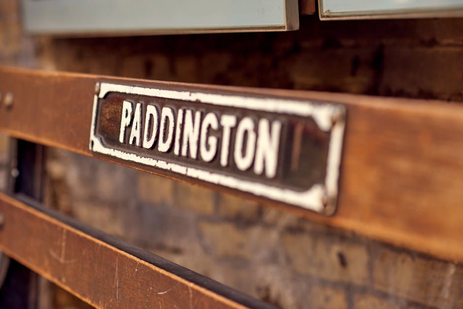 A close-up view of a vintage metal sign attached to a wooden surface, with the word 'PADDINGTON' prominently displayed in white capital letters on a black background. The sign has a weathered appearance with chipped paint and slight rusting around the edges, indicating age. It is positioned on what appears to be a wooden crate or storage box made of slatted dark brown wood, which is slightly blurred in the background. The setting suggests a storage or packing environment related to house removals or furniture transport, with natural lighting illuminating the textured surfaces. This image reflects elements of packing and moving logistics, suitable for content about home relocation or furniture transport services, as provided by companies like Paddington Man and Van.