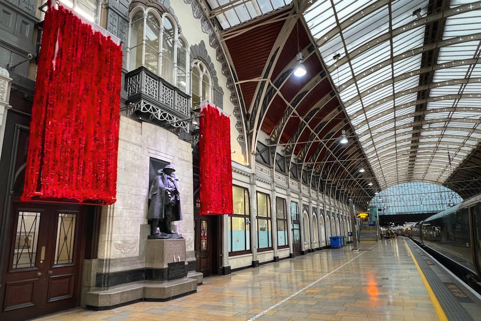 Inside Paddington Station, the image shows a spacious, well-lit platform with an arched, glass-paneled roof supported by metal beams. On the left side, there is a decorative façade featuring large red floral arrangements hanging vertically. Below these arrangements, a bronze statue of a man in period clothing stands on a pedestal next to a beige stone wall with ornate black wrought iron balcony details above. The platform has a wooden and tiled floor, with blue waste bins visible along the wall. The right side of the image displays part of a modern train with closed doors, ready for departure or arrival, and the platform stretches into the distance under bright overhead lighting. This setting, captured during daytime, illustrates the environment where professionals like Paddington Man and Van might carry out home relocation or furniture transport, including loading or unloading processes, in a safe and organized manner inside a prominent railway station.