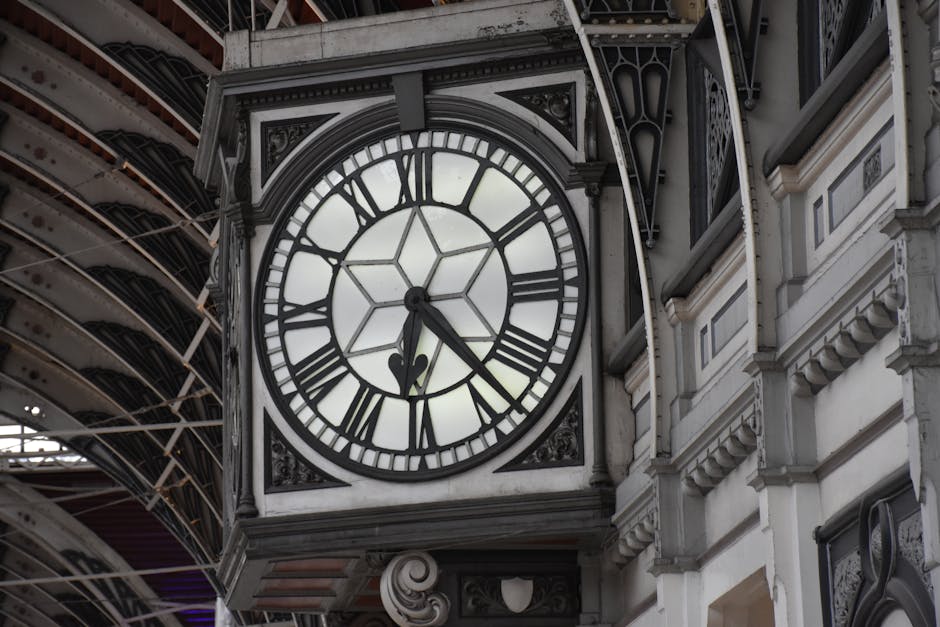 Close-up view of a large, traditional clock face with black Roman numerals and hands, mounted on the exterior of a historic building, with intricate architectural detailing and white stonework. The clock shows the time as approximately 10:26, and the background reveals a partial view of a covered walkway or train station with metal and glass roofing structure. The scene is well-lit with natural daylight, highlighting the ornate design elements and the clock's glass and metal frame. This setting reflects a typical urban location suitable for home relocation or moving services, such as those offered by Paddington Man and Van, with an emphasis on the environment where furniture or belongings might be loaded or unloaded during the moving process.