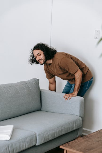 A man with curly black hair, wearing a brown T-shirt and blue jeans, is bending over to lift or reposition a light grey fabric-upholstered sofa inside a residential living room. The sofa is positioned against a plain white wall, with a small white towel or cloth folded on its armrest. To the right of the sofa, there is a wooden side table, and in the background, part of a white electrical outlet is visible. The scene appears to be part of a home relocation or furniture moving process, with the man possibly preparing to move the sofa into or out of the space. The room is well-lit with natural or indoor lighting. The image is associated with home removals and furniture transport services provided by Paddington Man and Van.