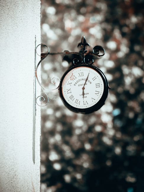 A round, black and white outdoor clock mounted on a decorative metal bracket attached to a light-colored exterior wall, showing Roman numerals and the words 'Reading Station' and 'London.' The clock is positioned on the left side of the image, with visible shadows cast by the bracket onto the wall. The background is blurred with a mix of brown, beige, and grey tones, suggesting trees or foliage. This scene is outside a property, possibly near a street or garden, representing a typical setting where house removals or packing and moving services, like those offered by Paddington Man and Van, may be relevant during home relocations or clearance activities.