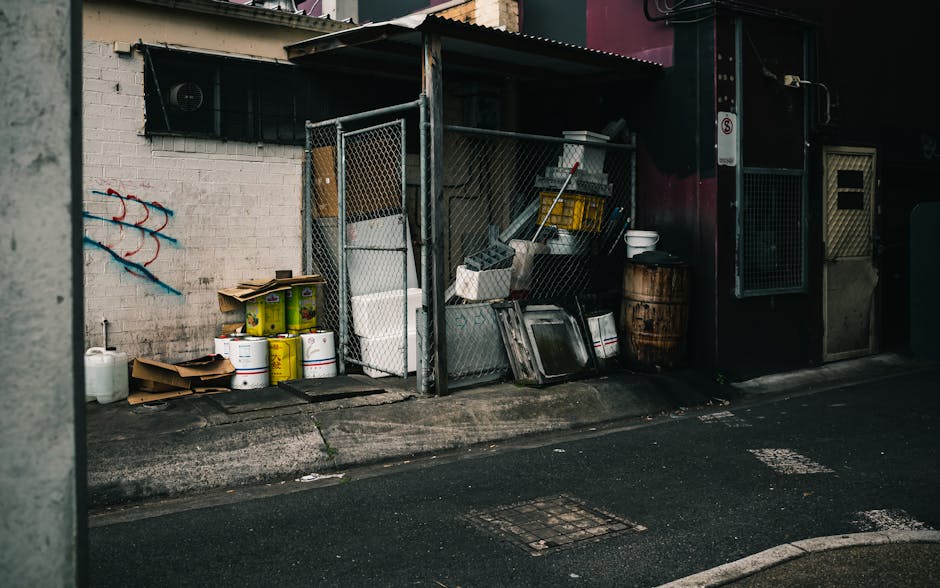 An outdoor area adjacent to a building features a small, open-sided metal storage shed with a corrugated roof, containing various items used in house removals and furniture transport. Outside the shed on the pavement are several cardboard boxes, plastic containers, and metal tins, some stacked and others placed individually. There are also large, white plastic buckets, a wooden barrel, and a few metal trays leaning against the shed walls. A white foam panel and a broom are visible within the storage area. The scene is illuminated by natural light, highlighting the urban surroundings including a white brick wall with some painted graffiti on the left side and a dark red painted section of the building to the right. The position suggests ongoing packing, loading, or disposal activities related to home relocation or bulky waste collection, with Paddington Man and Van's removal services likely involved in transferring these items onto a van, possibly for disposal or further transport.
