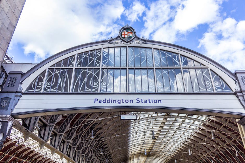 A clear view of the entrance to Paddington Station featuring an ornate metal and glass archway with decorative scrollwork and a crest at the top. The station's name, 'Paddington Station,' is displayed in bold blue text on a white sign beneath the arch. The lower section shows the loading bay area with a covered roof made of glass panels supported by metal framing, where furniture, cardboard boxes, and packing materials can be seen being prepared for loading. The scene is illuminated by natural daylight, with a partly cloudy sky reflected in the glass and visible in the background, illustrating the environment where house removals and furniture transport are carried out by professionals such as Paddington Man and Van, within the context of house removals, packing, and moving logistics.