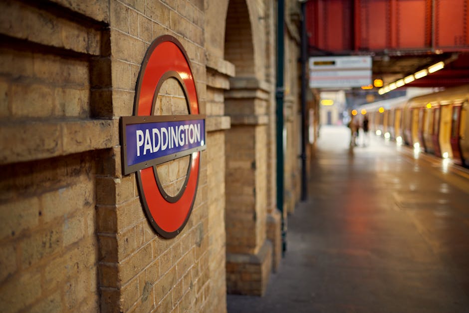 Close-up view of the Paddington underground station sign mounted on a brick wall, featuring the iconic red and blue roundel with white text, with the station platform and yellow train carriage visible in the background. The platform is under warm lighting, with a few blurred figures and luggage in the distance, illustrating an environment where home relocation projects involving furniture transport, packing, and loading processes might take place. The setting reflects urban train station surroundings in Paddington, part of a typical scene during house removals or moving services, with the station serving as a key transit point for transporting household items via train. The image emphasizes the infrastructure facilitating the movement of boxed belongings, furniture, and appliances in preparation for property transfer, supported subtly by [COMPANY_NAME]’s expertise in removals across the W2 postcode area. The lighting highlights the textures of the brickwork and station signage, capturing the typical ambiance of a busy, functioning transport hub ideal for supporting detailed logistics planning in home relocation efforts.