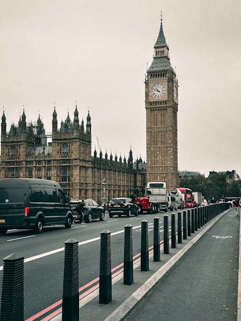 A view of the Palace of Westminster in London, with the iconic clock tower commonly known as Big Ben prominently visible. The image shows a busy street scene with multiple vehicles, including black vans, white buses, and private cars, queued along the road. In the foreground, a designated cycle lane is separated from the main traffic by black plastic bollards. The road appears to be part of an urban setting, with the historic Gothic-style architecture of the Palace of Westminster in the background under an overcast sky. The scene captures the typical city transport environment, where vehicle and pedestrian movement coexist, and reflects ongoing urban activity related to city logistics. Paddington Man and Van, a professional removals company, could use this setting as part of their home relocation and furniture transport services, illustrating the busy street context for loading or delivering household items during a house move.