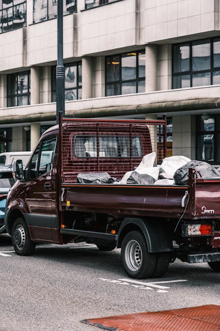 A small burgundy flatbed cargo van parked on a city street in front of a modern office building with large windows and concrete facade. The van's cargo area is filled with wrapped furniture pieces and moving boxes covered in plastic or fabric, indicating a home relocation or furniture transport process. The loaded items include wooden furniture, cardboard boxes, and soft coverings, secured with straps. A metal safety grate is fitted at the back of the van to protect the cargo during transit. The surrounding environment features other parked vehicles, a streetlamp, and a section of red-brick pavement in the foreground. Natural daylight illuminates the scene, highlighting the details of the vehicle and the packed contents, which are prepared for loading or unloading. This image showcases a typical scene of professional packing and moving logistics, relevant to house removals and same-day emergency relocations for Paddington clients, as offered by Paddington Man and Van.