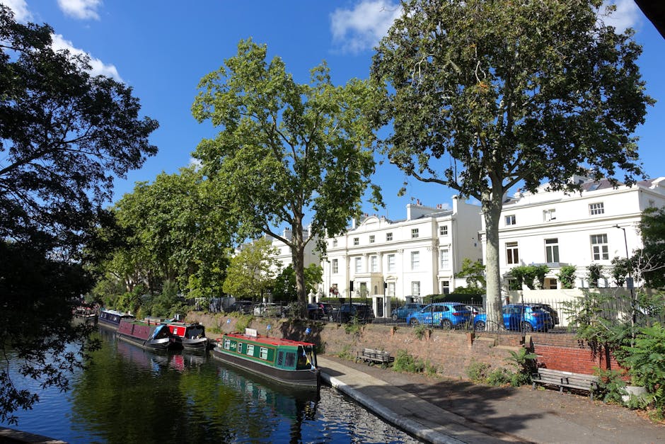 A sunny daytime scene along the canal at Sussex Gardens shows several houseboats moored side by side, with their exteriors painted in various colors, including dark green, maroon, and black. The boats are positioned parallel to the canal edge, which is lined with a narrow walkway made of concrete and brick, bordered by a metal railing. Behind the walkway, tall leafy trees with lush green foliage cast shade over a row of white Victorian-style terraced houses with large windows, decorative cornices, and small front gardens enclosed by wrought iron fences. Parked cars in shades of blue and grey are visible along the street adjacent to the houses. The sky is bright blue with a few white clouds, and the overall scene captures a peaceful residential area typical of London’s Paddington district, as seen in images related to house removals and relocation services by Paddington Man and Van.